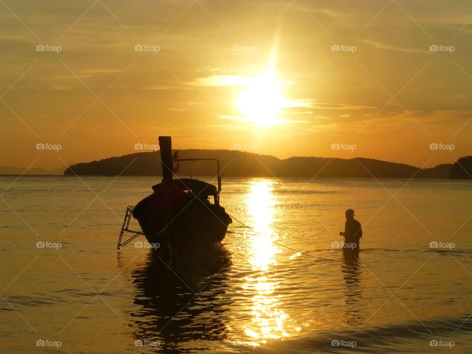 A man walking into the sea towards a longtail boat at sunset