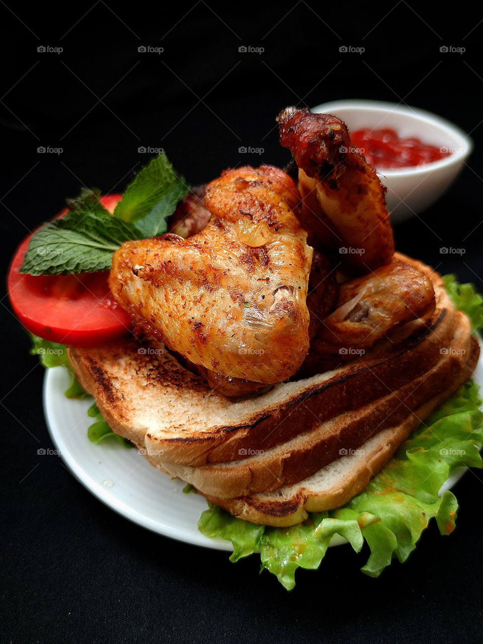 on a white plate: green lettuce leaves, croutons with pieces of fried chicken wing, a piece of red tomato with a green mint leaf on it.  In the background is a white gravy boat with red ketchup.  Black background