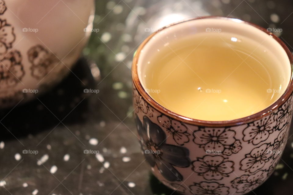 Close up of a small ceramic cup of Japanese green tea with a blurred background of the table and part of the kettle. 