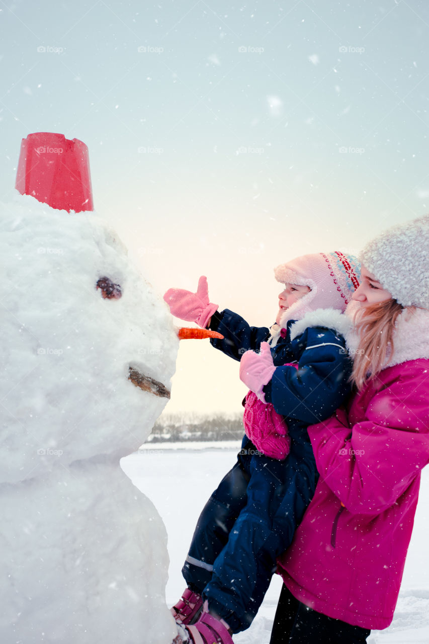 Girl and her little sister making a snowman, spending time together outdoors, having fun in wintertime