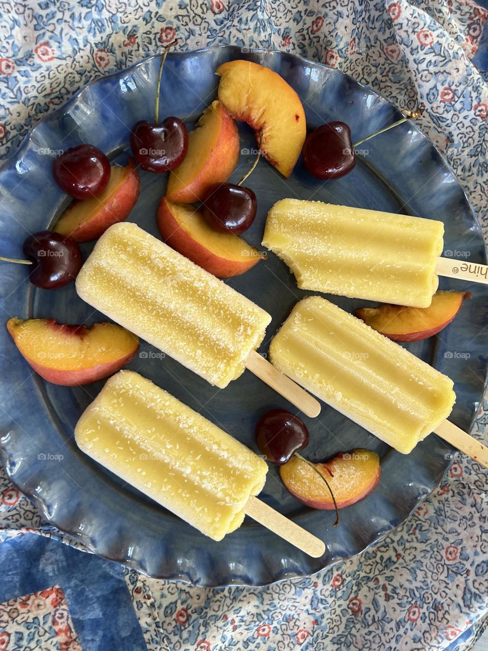 Four yellow pineapple popsicles with black cherries and nectarine slices on a blue ceramic plate placed on a blue and orange flower Liberty type printed scarf