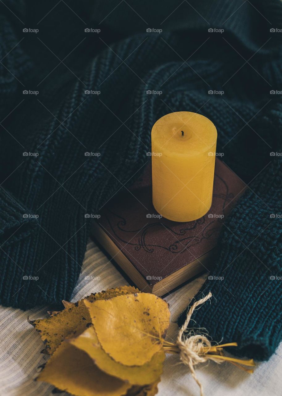One yellow candle, a book, a bouquet of autumn leaves and a warm knitted dark green sweater lie on a bed with a light blanket, close-up view from above.