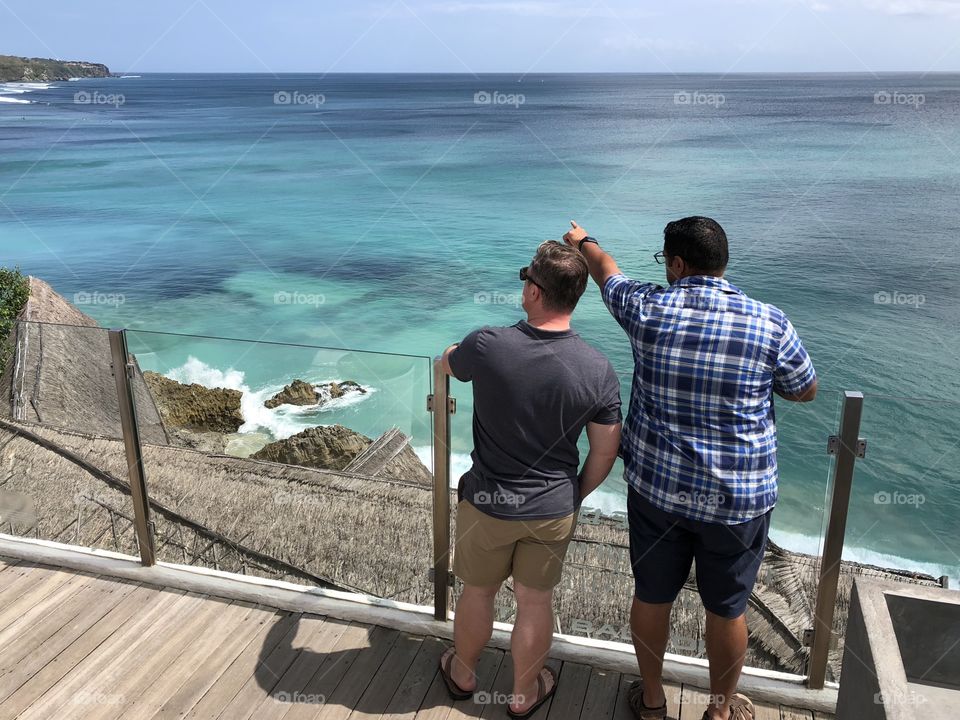 2 men at a cliffside spot, overlooking amazing blue ocean water. Pointing. Diverse. Bali. 2018.