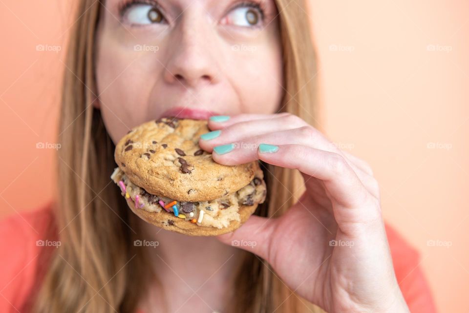 Fun product portrait shot of a woman taking a bite out of a large chocolate chip cookie sandwich 