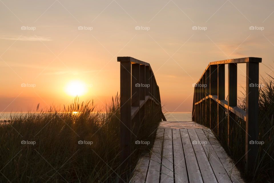 The sun is shining on a wooden boardwalk leading to the ocean at sunset on a warm summer evening 