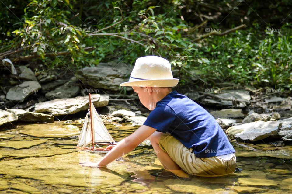 Young boy playing with a toy sailboat in a creek with the sunlight reflecting on the water 