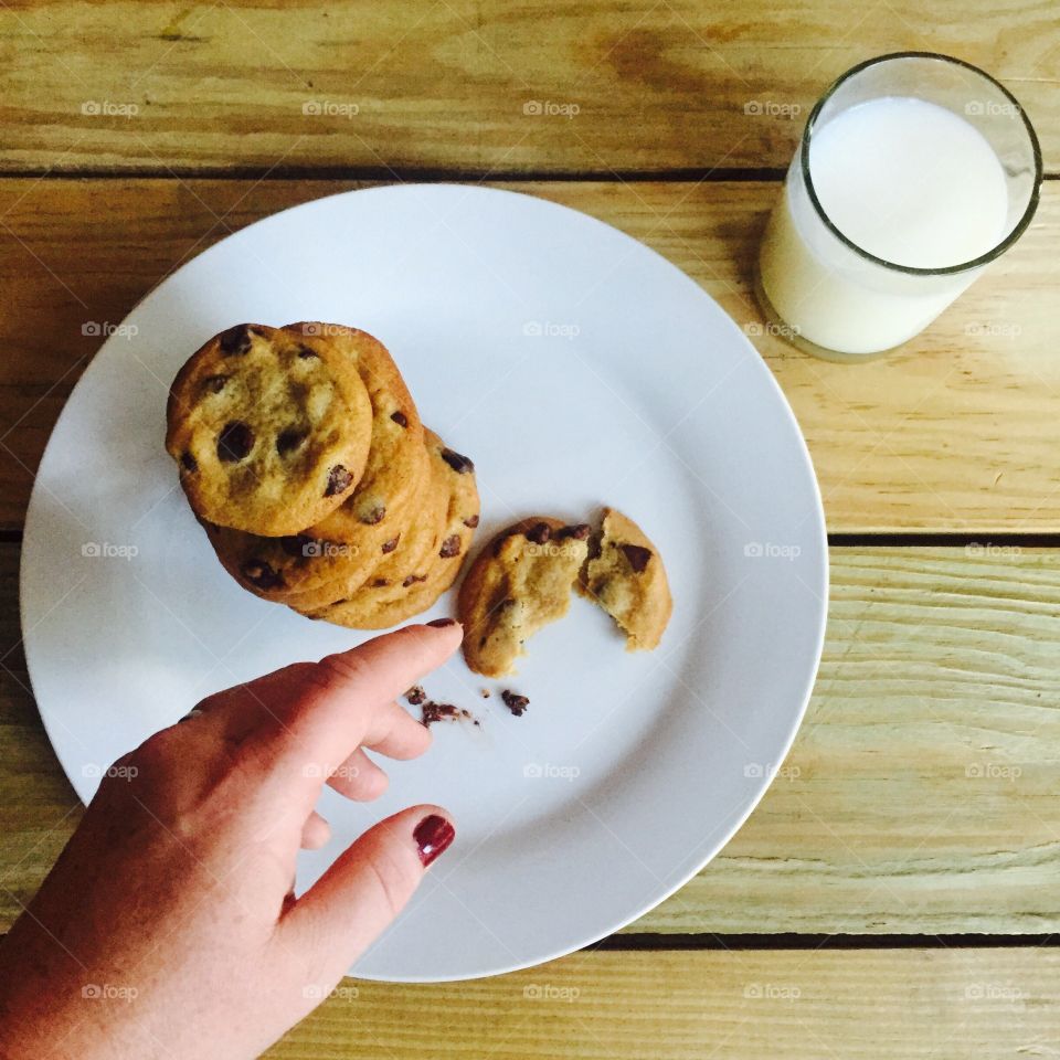 One Last Cookie. Woman's hand reaching for a cookie from a plate of chocolate chip cookies on a white plate with a glass of milk in view