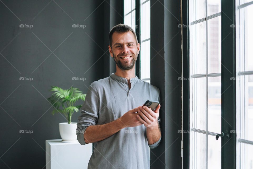 Young bearded man in gray longsleeve with smartphone in hands reading message standing at window in modern office with dark walls