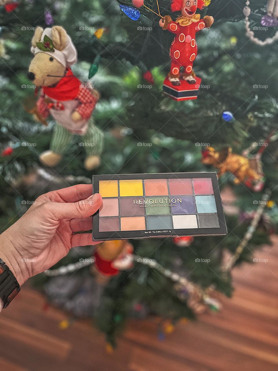Woman holding eye shadow palette by Christmas tree, woman putting a present under the tree, eye shadow palette Christmas gift, woman’s hand holding beauty product, beauty products for Christmas presents, colorful makeup products