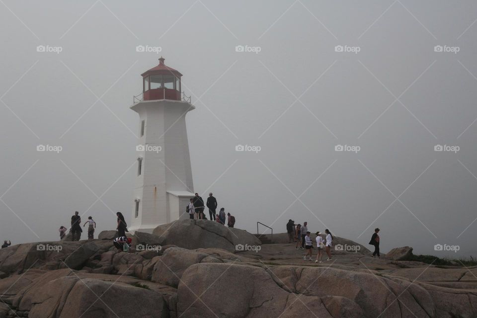 Crowds of tourists gathered on the rocks surrounding the Peggy's Cove lighthouse on a foggy afternoon.