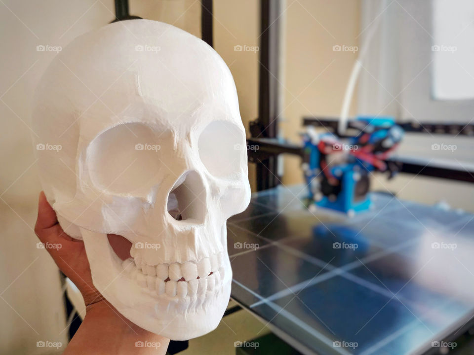 3D printed skull model on human hand with blurred background of 3D printer for copy space on the right side. Medical model part concept.