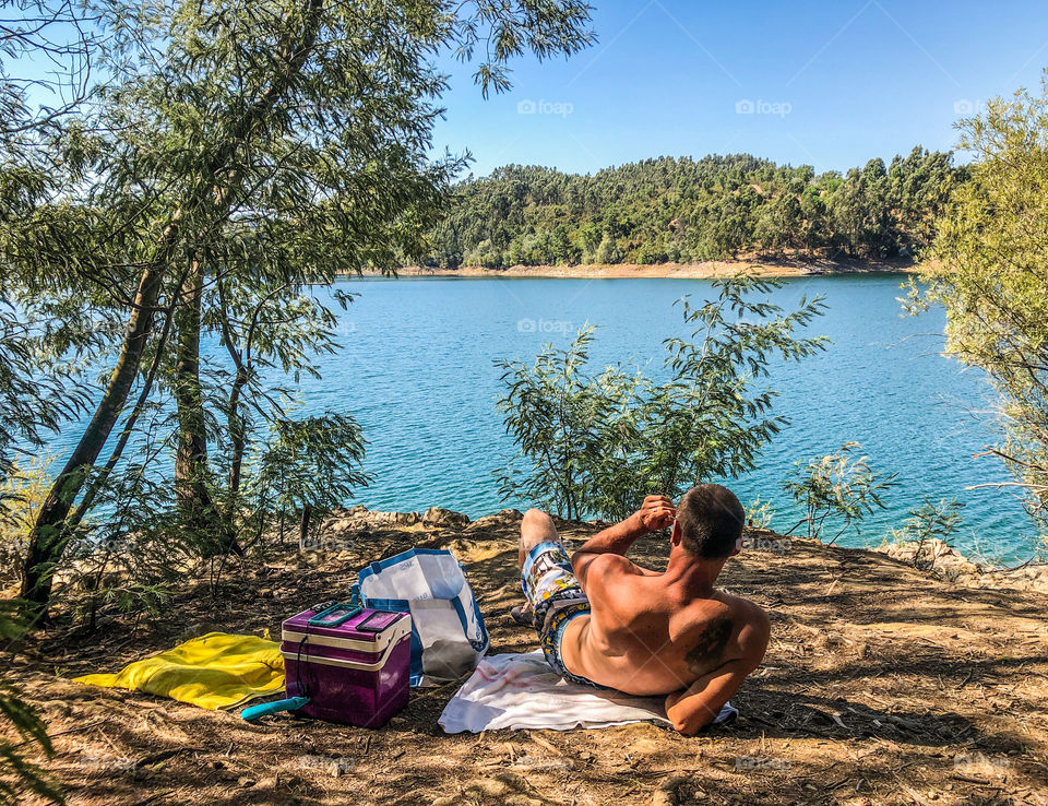 Tanned, young man sits relaxing and taking in the river view in Portugal. Towels, a picnic bag and cooler sit beside him