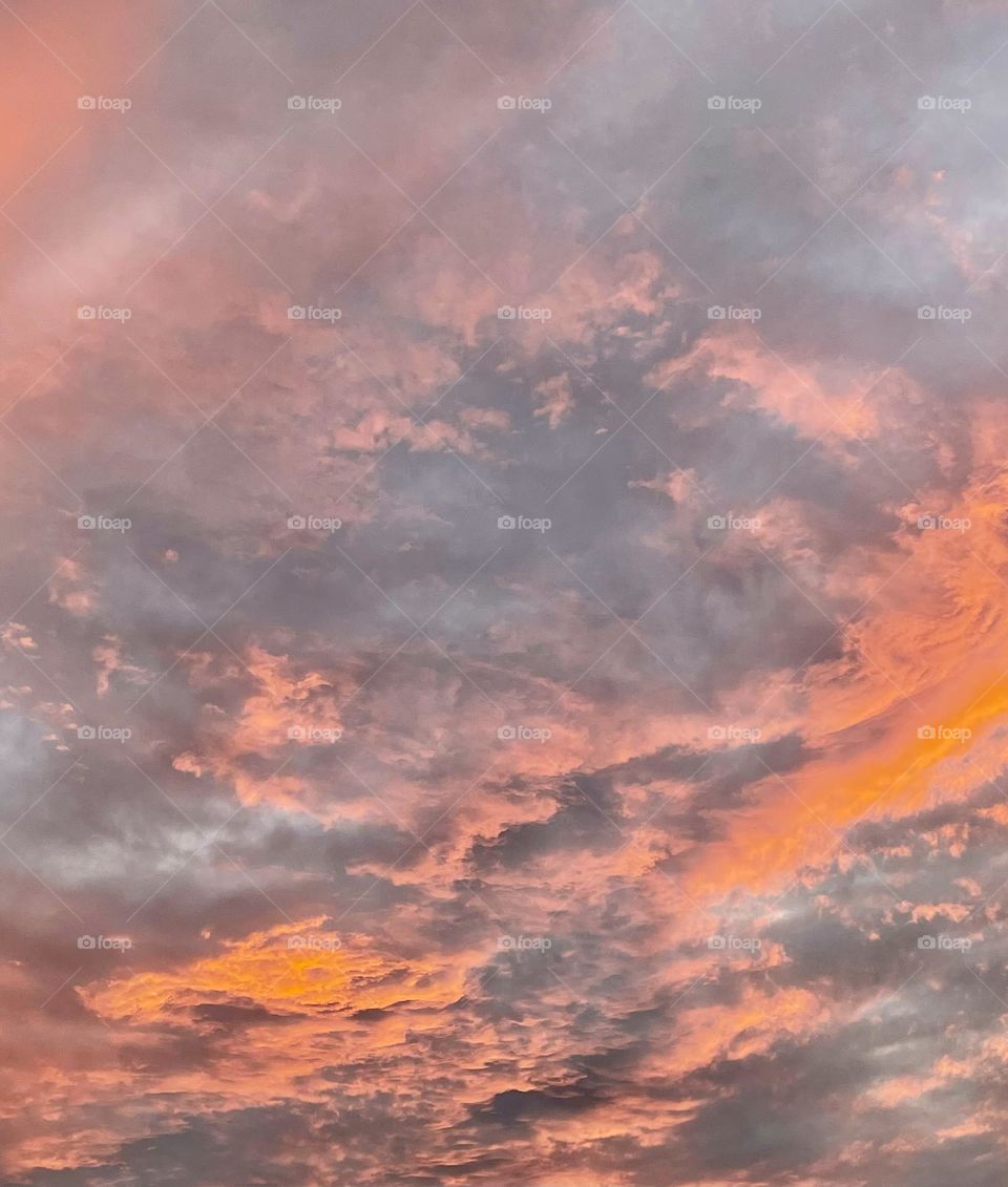 The summer evening sky unfolds like a vast, vibrant canvas, brushed with strokes of golden orange and pale red, that melt into deeper gray-blues, creating a dynamic yet harmonious gradient. The varying cloud texture adds depth to the beautiful sky.