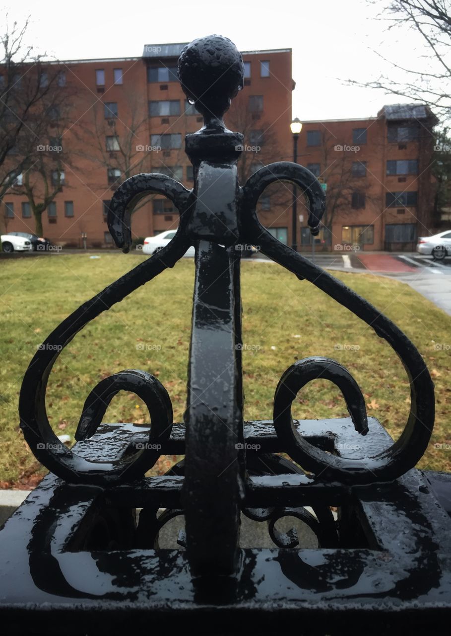 The curly rod iron top of a fence that is painted black. The day is gray and rainy. The fence is wet.