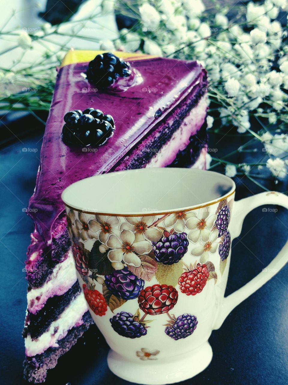 A mug with bright purple blackberries and red raspberries painted on it. Behind the mug is a piece of purple cream cake. Decorates the cake with purple blackberries