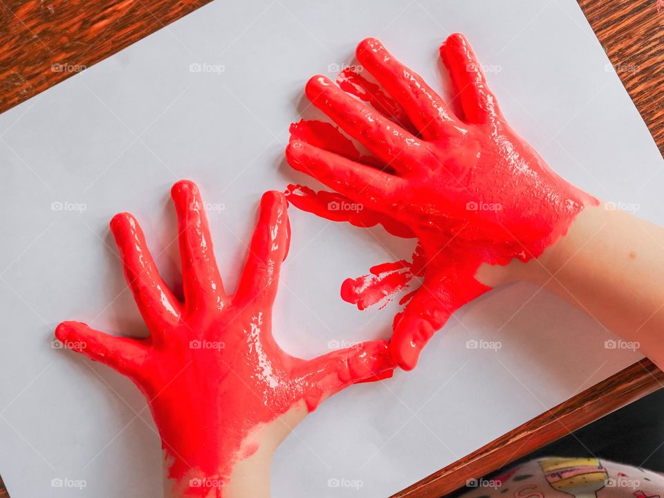 Little caucasian girl prints her hands painted with red paint on a white sheet, sitting at a wooden table, top view close-up. Kids creativity concept.
