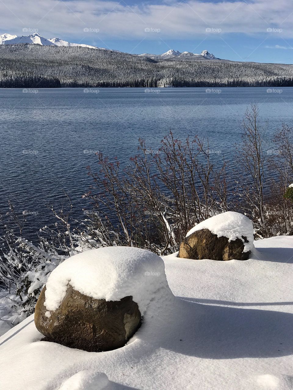 Two large snow covered boulders on the shores of the beautiful blue Crescent Lake in the mountains of Oregon with snow covered mountains in the background on a sunny winter day. 