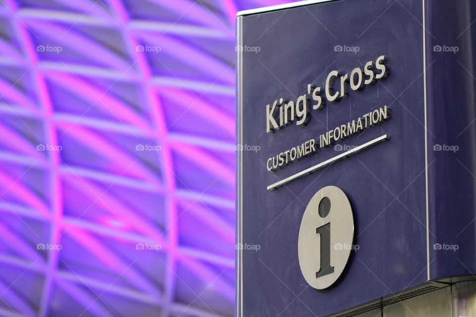 An informative sign at King's Cross train station in London denoting the location of a customer service help desk with the famous purple lattice roof in the background.