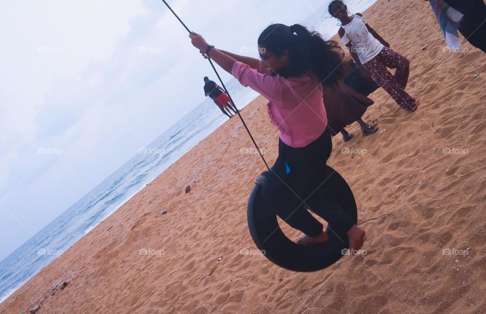 This is a teenage girl spending her leisure time very happily and funnily. She is swinging on a  swing made by a tire on the sea beach. She seems so funny and she is very happy There are some others near her and the blue blue ocean is staying beside.