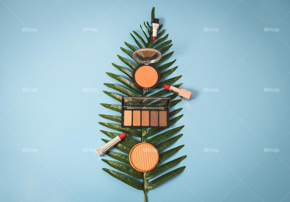 A nude eyeshadow palette, round face powder and three red lipsticks on a palm tree branch lie in the center on a blue background with a copy of the space on the sides, flat lay, close-up. Concept female cosmetics, beauty salon.