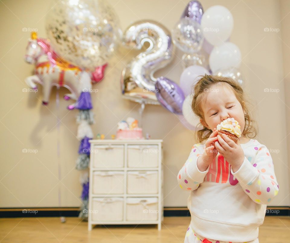 Small birthday girl in pajamas eat cupcake and close eyes in her room with balloon