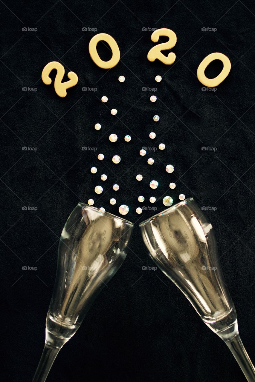 Flat lay of the numbers ‘2020’ in gold above gold-tinted champagne glasses with rhinestone bubbles on black background - portrait format