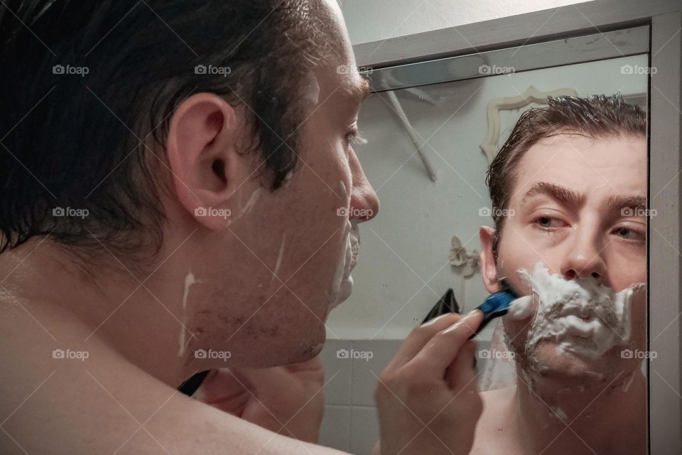 A man shaves while looking in the bathroom mirror