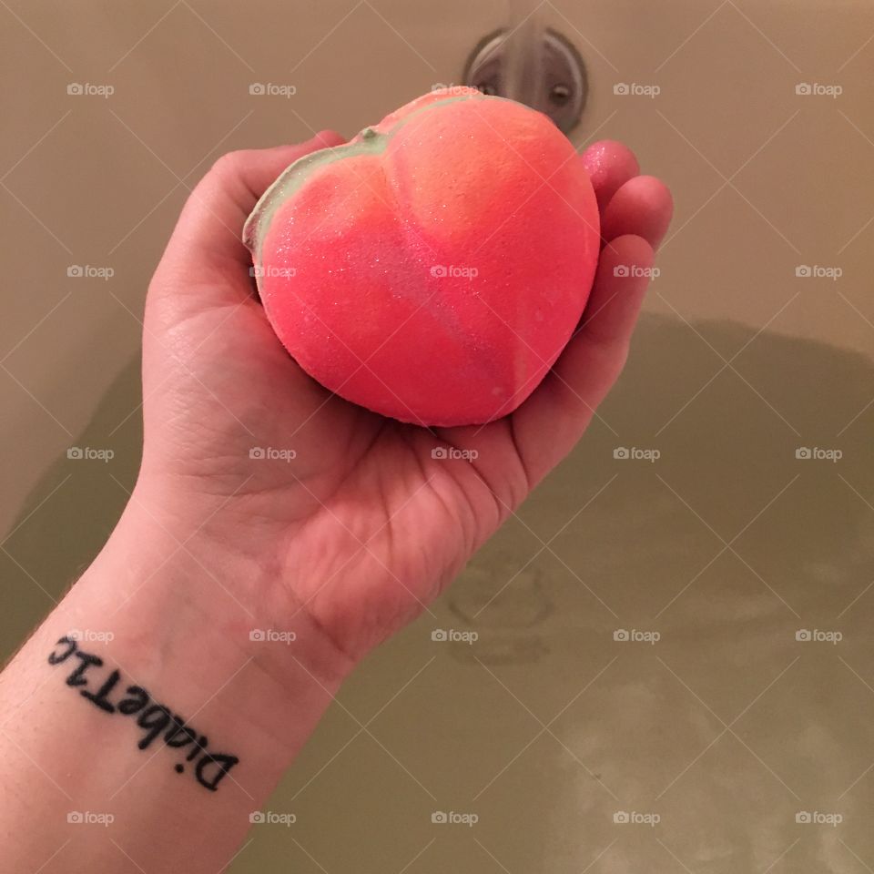 Hand holding a colourful peach shaped bath bomb. Arm has a diabetes tattoo. Running bath water in the background. 