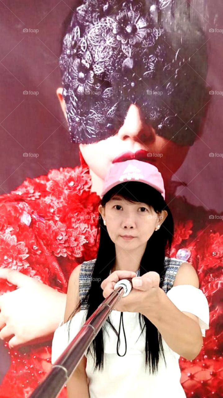 A woman wearing a white and gray shirt is taking a selcie usinģ a selfie stick in front of a poster