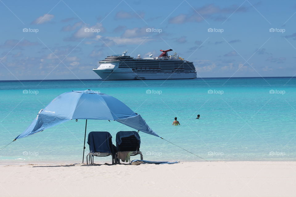 Umbrella with two lawn chairs and a couple in the water where is the cruise ship in background for vacation travel