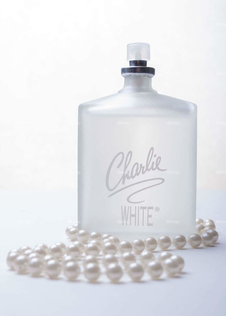 monochrome white on white bottle of Charlie White perfume surrounded by a string of white pearls on a white table in front of a white background