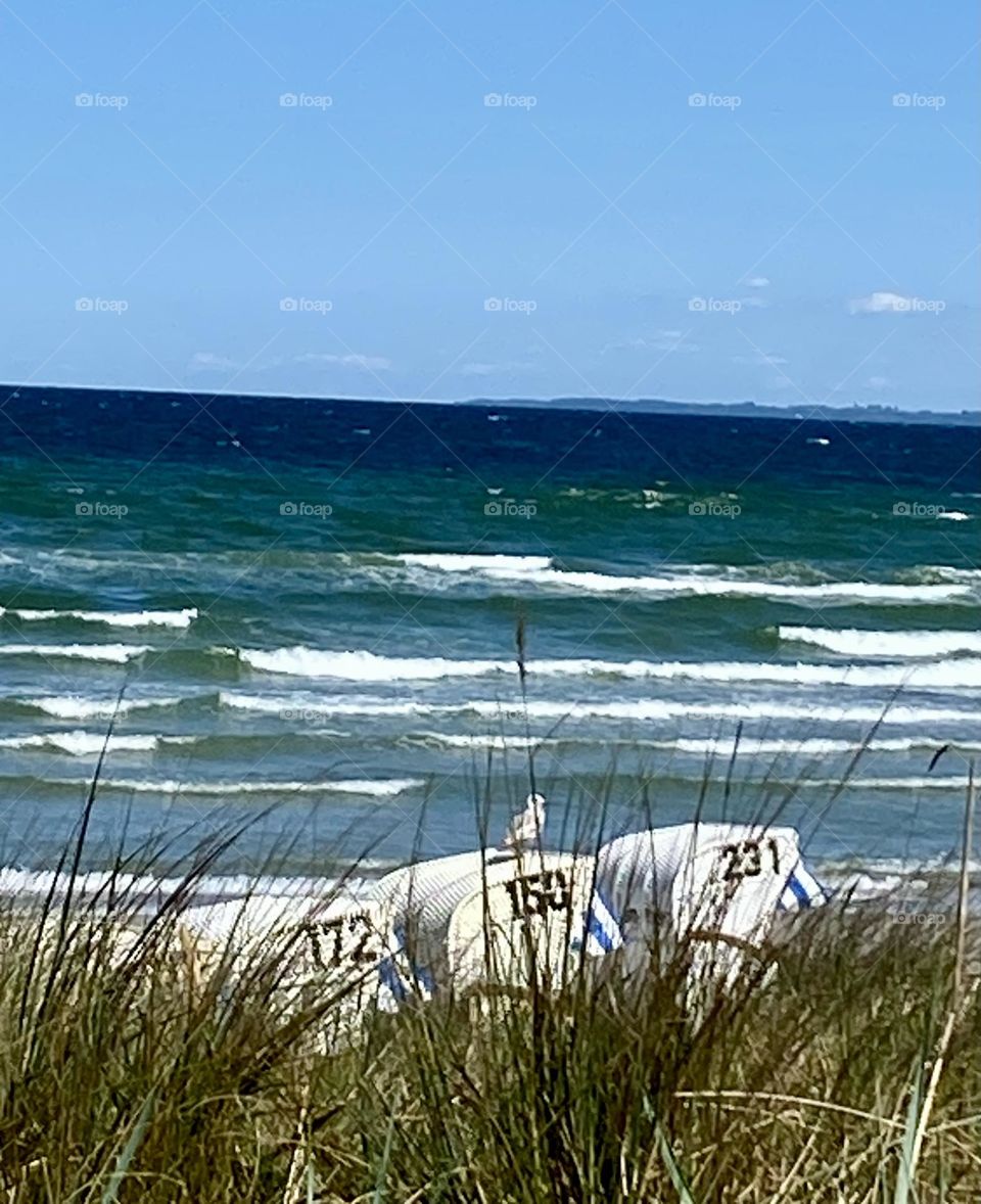 The deep blue sea ends with shallow waves on the beach. Three beach chairs are visible behind beach grass. A fresh breeze sets the picture in motion. A seagull sits on the middle beach chair.