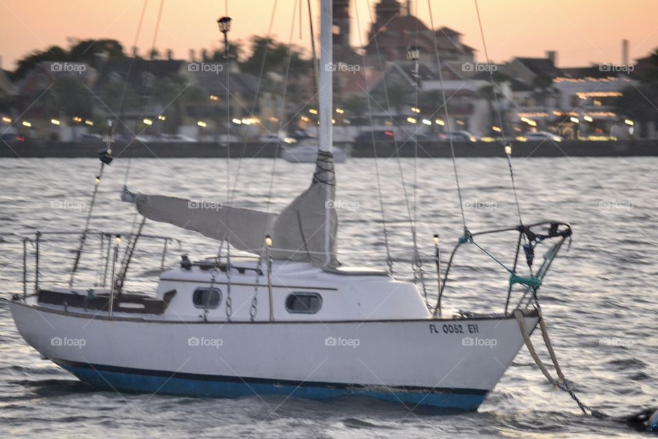 A closeup view of a white sailboat on a river with a city in the background and an orange sky at sunset