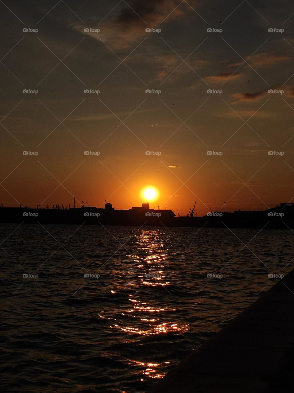 A photo of a sunset over the sea in Greece with beautiful colours of the sky and the machinery of the sea port visible in the back