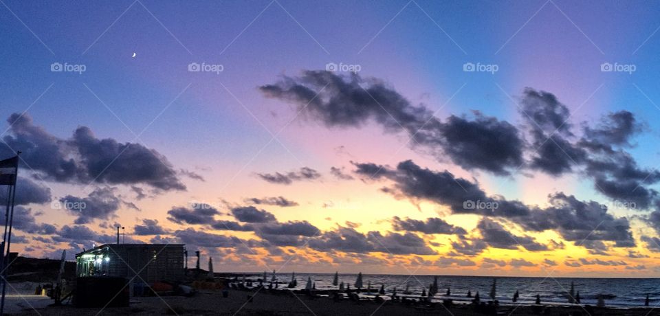 An amazing sunset sheds light in the sky while the moon rises up to show its presence. The beach bar switches on the indoor lights to get ready for the darkness.