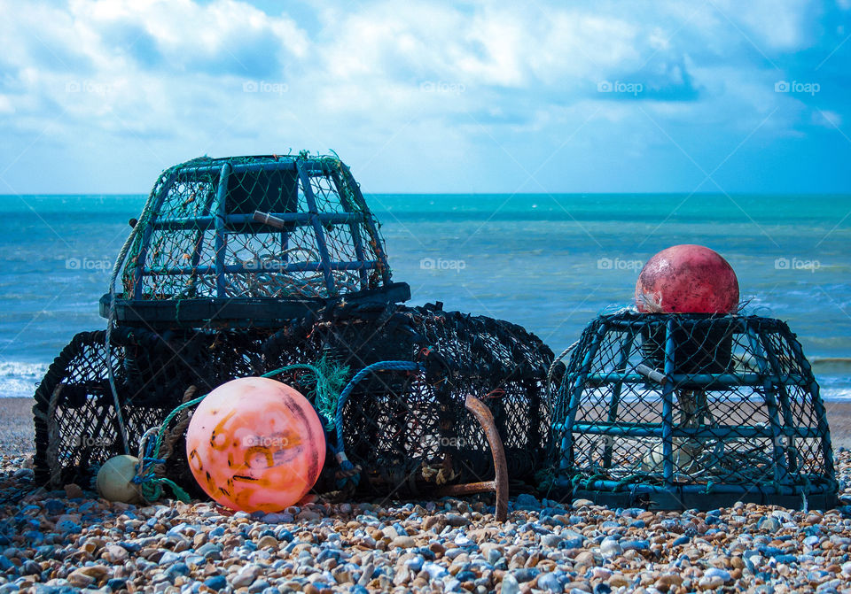 Orange buoys and blue lobster pots sit on a pebbled beach with blue sea and sky in the background 