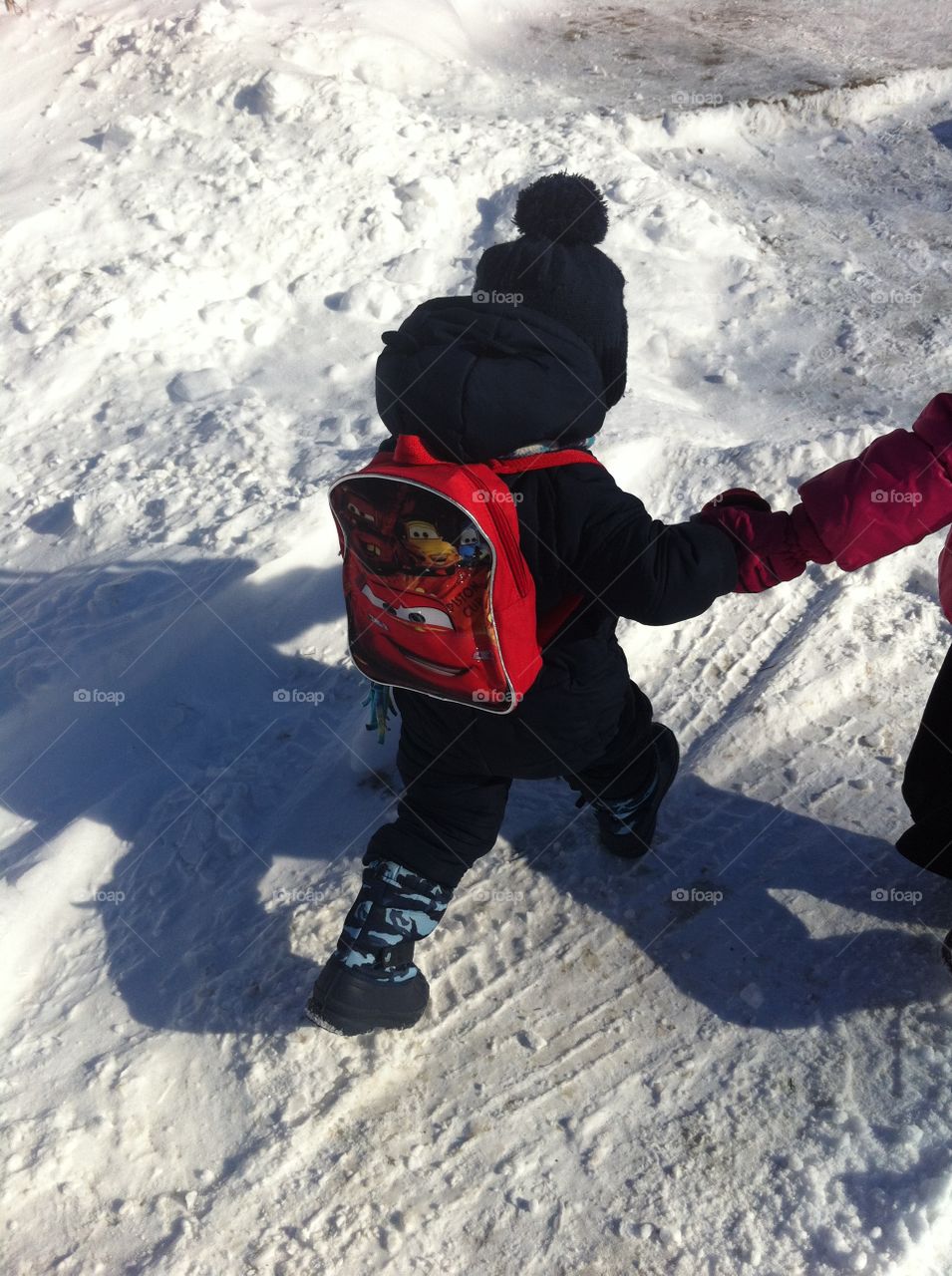 Toddler boy walking to school in the snow while holding hands, carrying a Cars backpack and wearing a touque, snowsuit and boots