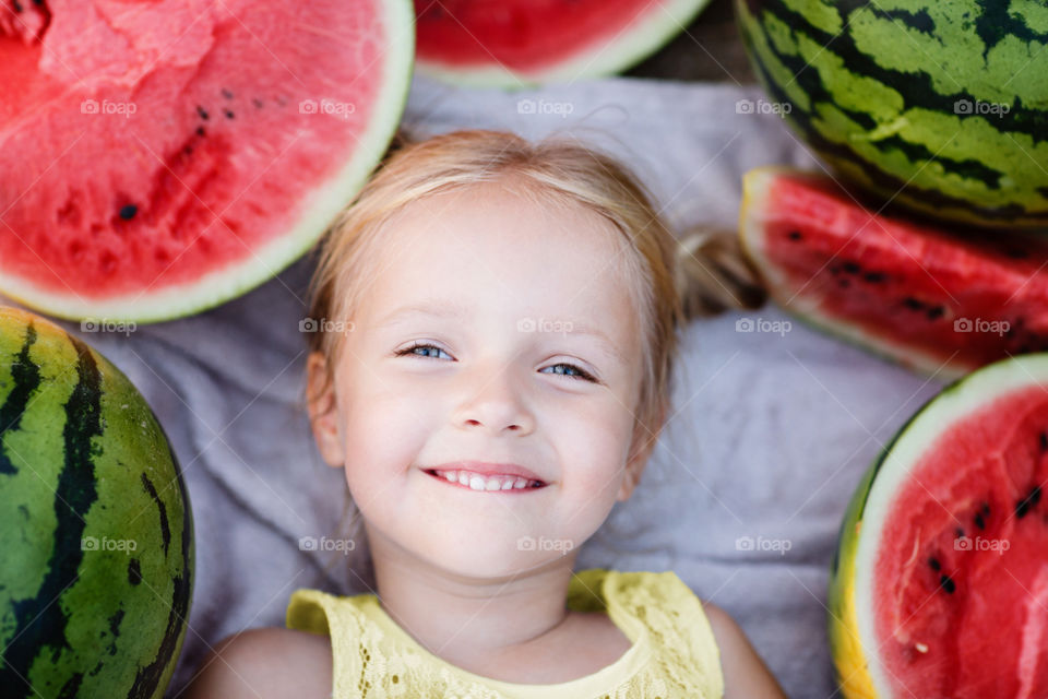Portrait of cute little girl with blonde hair lying on blanket with fresh watermelon 