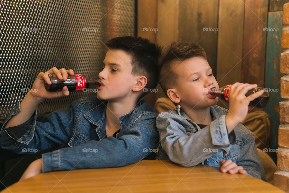 two lovely brothers, dressed in denim clothes, with beautiful hairstyles, are sitting in a cafe and drinking Coca-Cola. communicate, laugh, smile. two guys, two friends