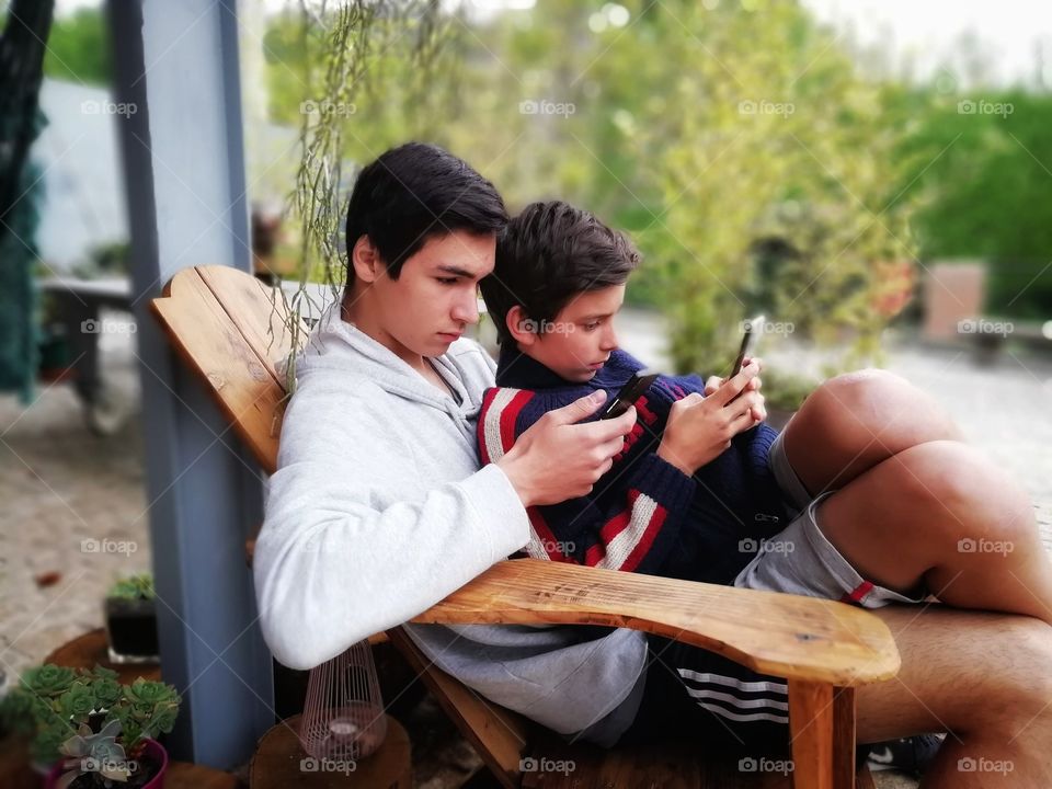 A teenager and a young boy sitting together on a wooden chair, on an outdoor terrace, surrounded by nature on a spring afternoon, super concentrated on their mobile phones