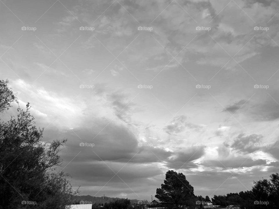 A black and white photo of a cloudy sky with trees and mountains far in the distance. 