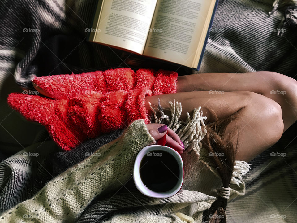 Overheard view of a young woman in cozy pink home boots reading a book and drinking coffee in cozy bed 