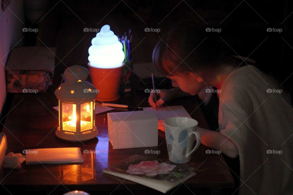 Little girl writes at the table by the light of a table lamp and a candle