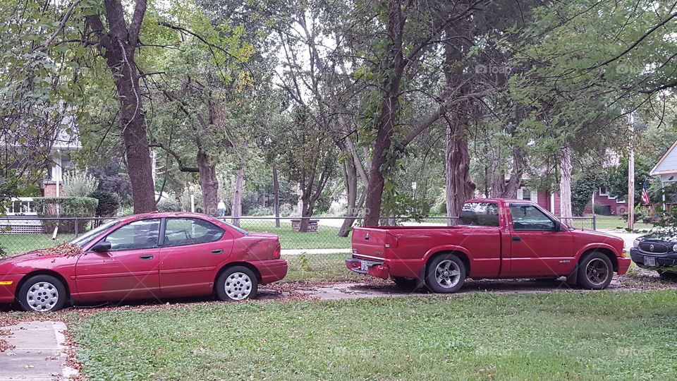 A red car and a red truck in the driveway. Tons of trees in the background and the white dot is my dad walking our dog.