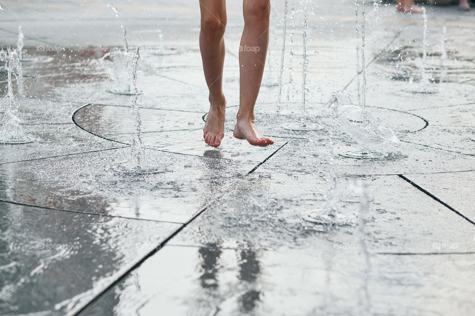 Child standing, jumping, playing in a fountain while hot weather. Closeup of legs, water streams, splashes. Candid people, real moments, authentic situations