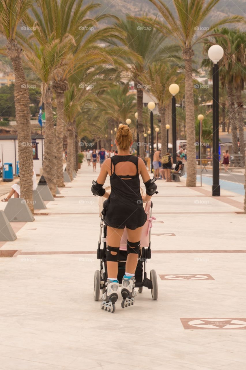 A woman combining sports with taking a walk with her kids by skating on the boulevard behind the stroller in alfaz del pi, spain