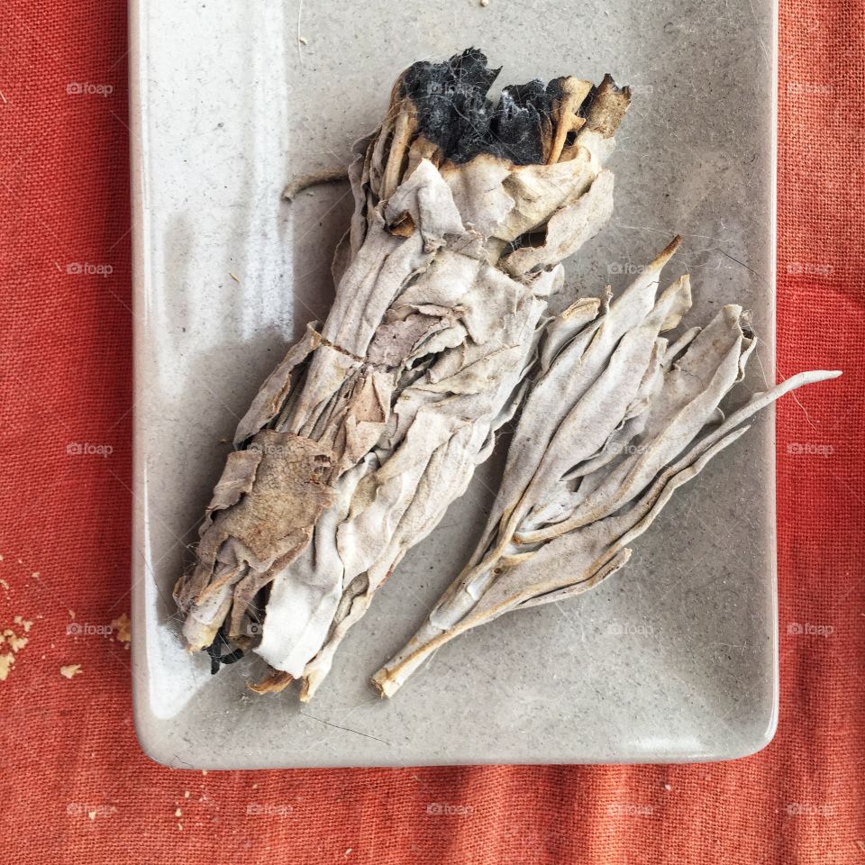 A half burned sage smudge in a dish on a red cloth background. The smudge is accompanied by a smaller set of dried sage leaves.