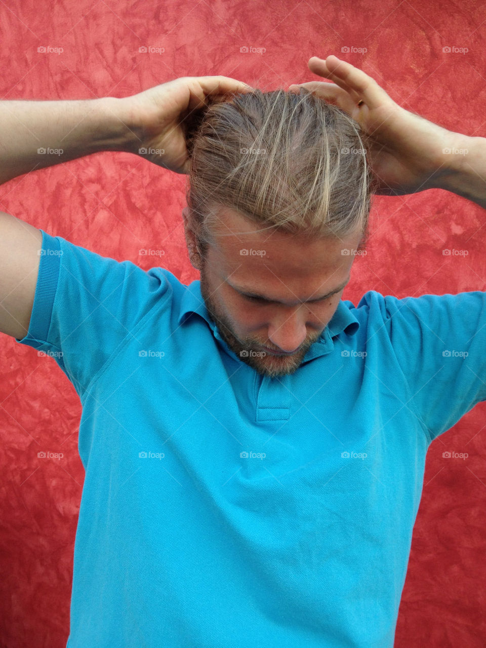 Man with long hair. Man with long hair in turquoise shirt is putting his hair together in a ponytail and stands in front of a red wall
