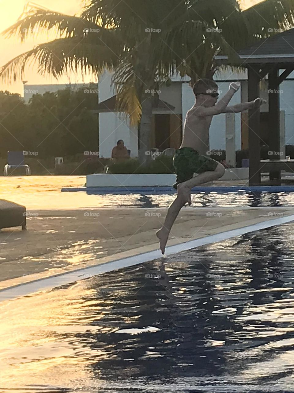Boy jumping in to a beautiful pool with the golden sun setting behind him and palm trees above him.