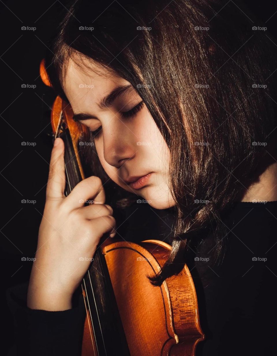Little girl in black, holding a violin in her hand on a black background and the rays of the sun fall on her